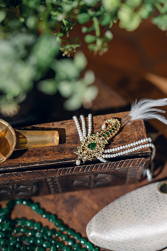 Groom accessories flatlay with groom jewelry details, pearl and green gemstone brooch, pearl strands, feather, jutti shoes on wood tabletop with greenery