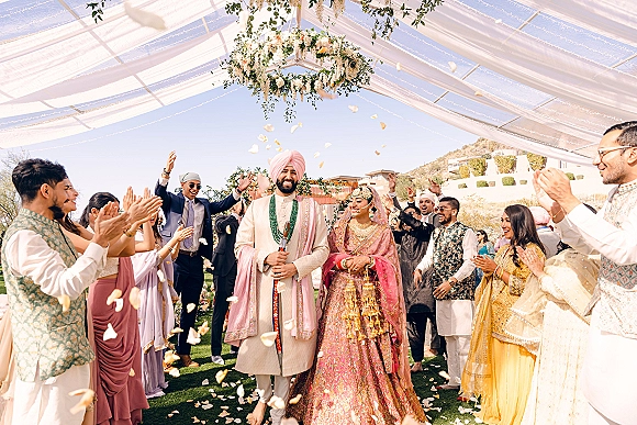 Wedding recessional as bride and groom walk the aisle during a rose petal toss under a draped canopy with hanging greenery and lights outdoors
