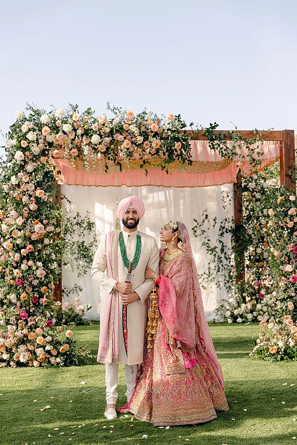 Couple portrait of a Sikh wedding couple under a rose and greenery floral arch with draped fabric on an outdoor lawn under sky