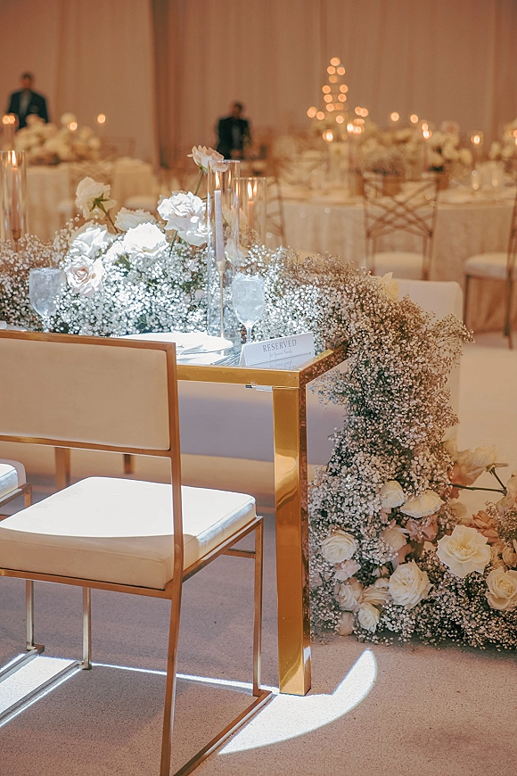 Wedding sweetheart table with white roses and baby's breath garland, taper candles and reserved sign on a gold mirrored table before draped backdrop