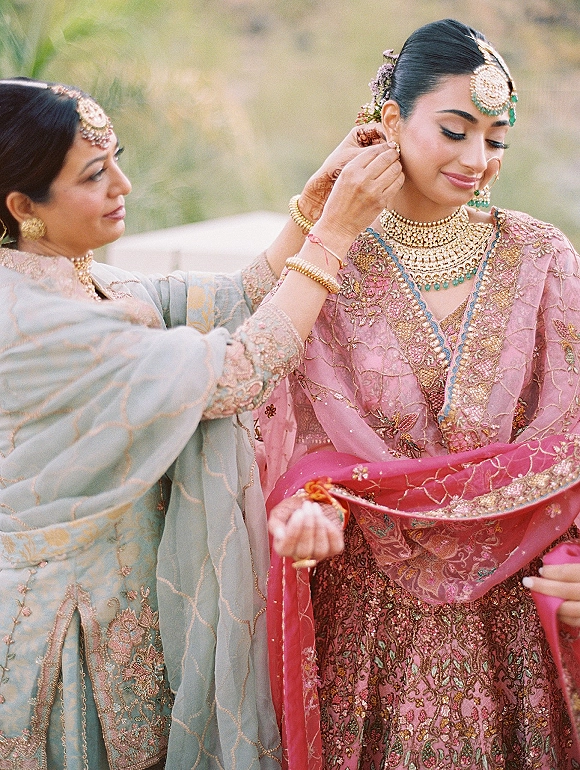 Bride getting ready as her mother fastens chandelier earrings, wearing a pink embroidered lehenga and dupatta, with greenery behind