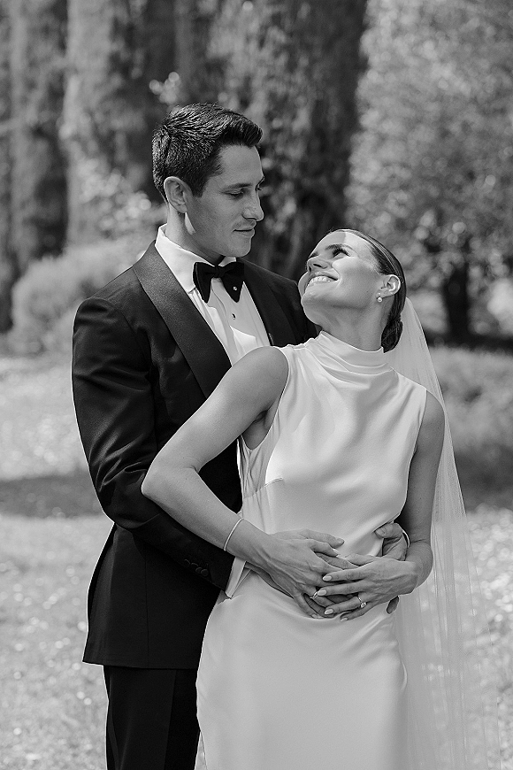Couple portrait in black and white, groom hugging bride from behind as she looks up at him on a garden pathway with trees