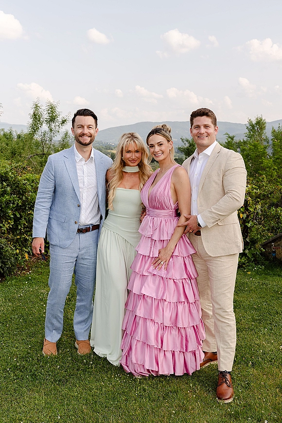 Wedding guest group photo of friends in pastel dresses and light suits, with a pink ruffle gown on a grassy lawn and mountains behind