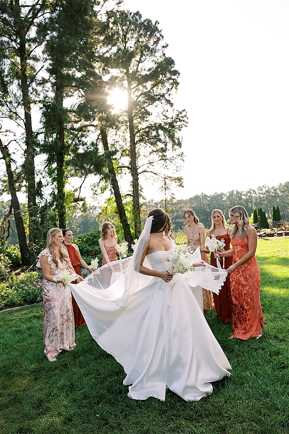 Bride with bridesmaids in rust dresses holding her long train and bouquets, cathedral veil trailing on a sunlit lawn under tall trees
