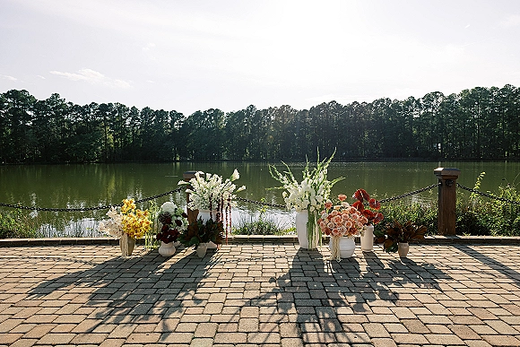 Ceremony floral decor with ground floral arrangements in white vases, peach roses and colorful blooms lining a brick patio by a lake