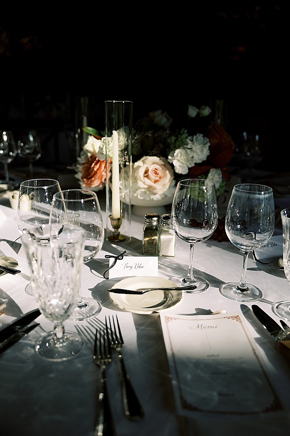 Reception tablescape with a wedding place setting, crystal glassware, menu and place card, and rose centerpiece with taper candle in a dim room