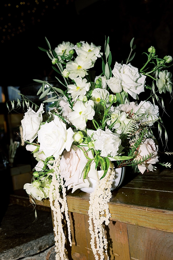 Wedding floral arrangement of white and blush roses with greenery and trailing amaranthus in a ceramic vase on a wooden table, bistro lights behind