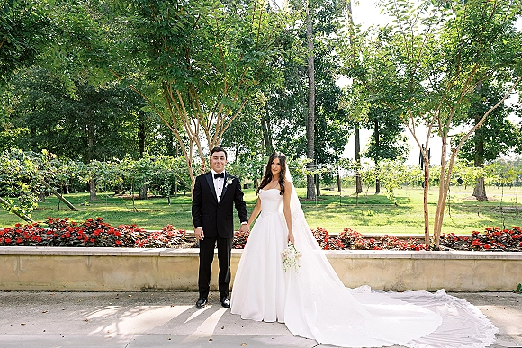Couple portrait of bride and groom holding hands, her veil and long train flowing beside a bouquet on a garden walkway near a stone wall