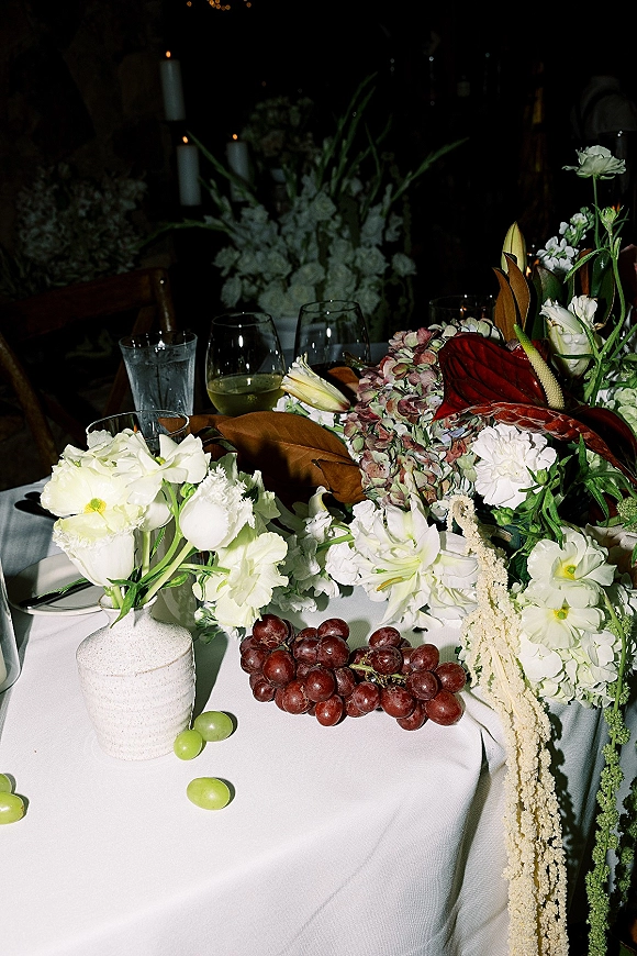 Reception tablescape with wedding table centerpiece in a ceramic vase, white hydrangea and anthurium, taper candles, grapes, and wine glasses in candlelit room