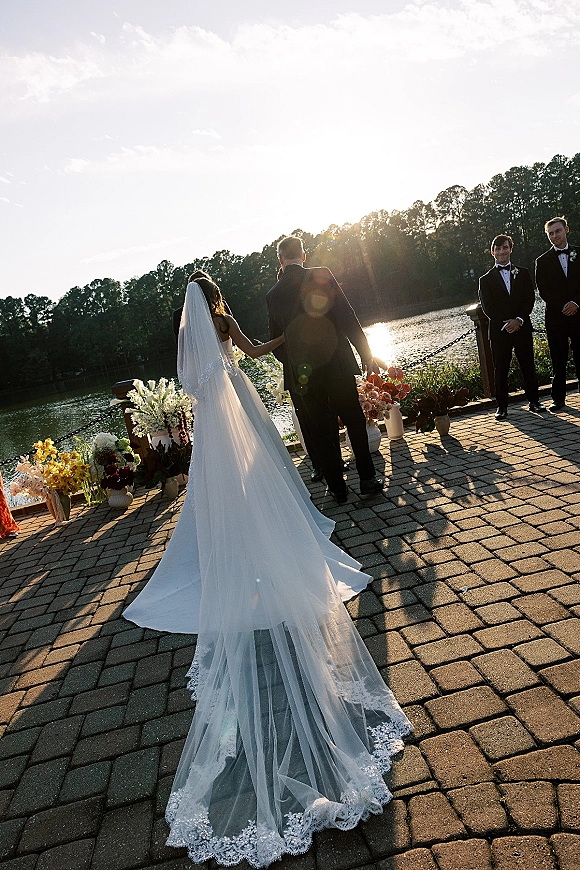 Wedding processional as bride walking down aisle with veil and long train, groomsmen in tuxedos on a lakeside patio backdrop