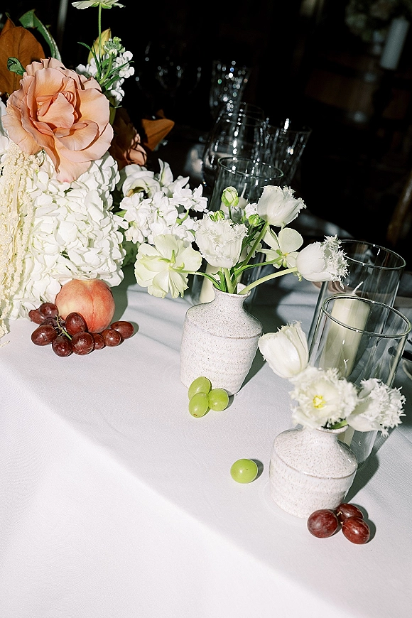 Reception tablescape with wedding table flowers in ceramic vases, white hydrangea and peach roses, candles, glassware, grapes in a dim room