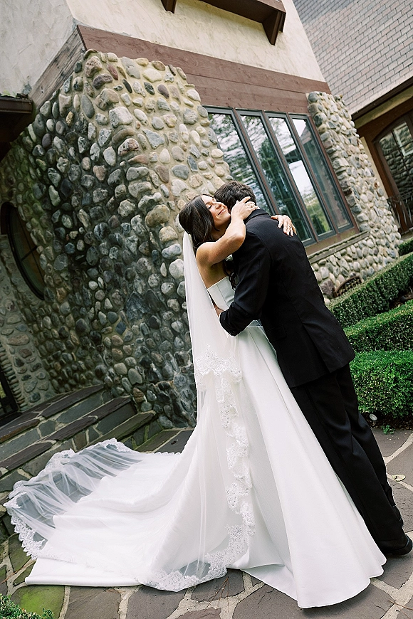 Wedding couple portrait of bride and groom hug, her long lace-trim veil and dress train flowing on a stone walkway by windows