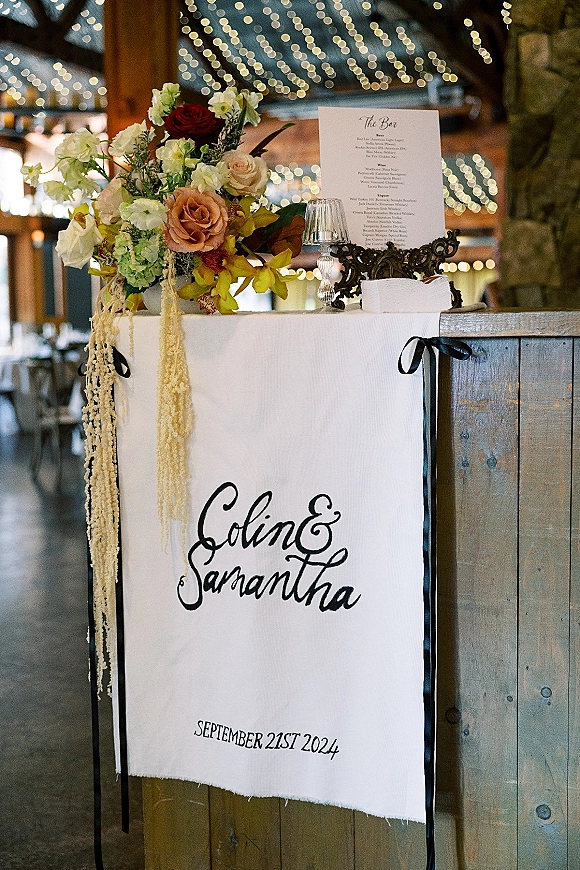 Wedding bar sign with signature cocktail menu in black calligraphy on fabric, framed by roses and orchids on a rustic indoor bar under string lights