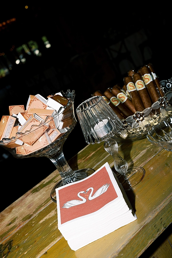 Wedding favor table with cigar bar wedding details, matchboxes and glass bowls on rustic wood under bistro lights in a dark reception space