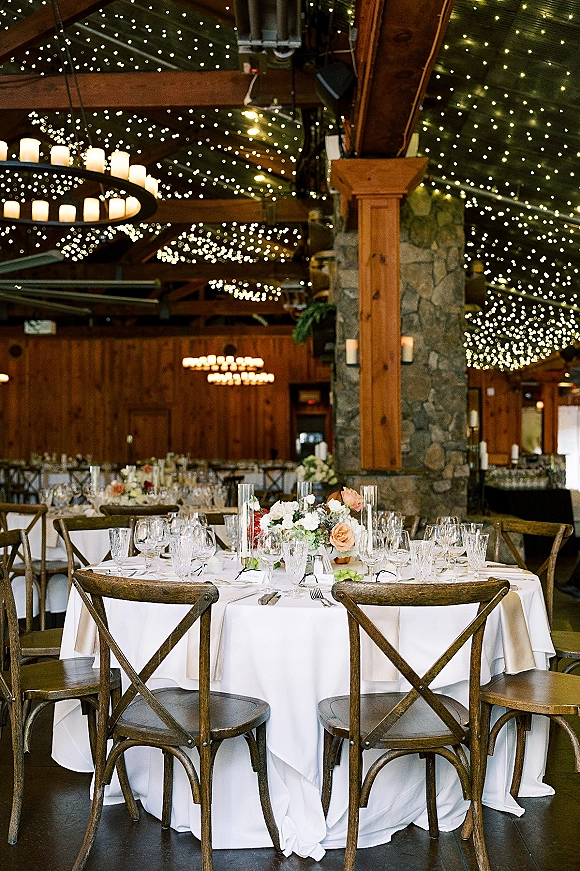 Reception tablescape with round wedding table setup, white linens, floral centerpiece and candles under chandeliers and string lights in a wood barn interior