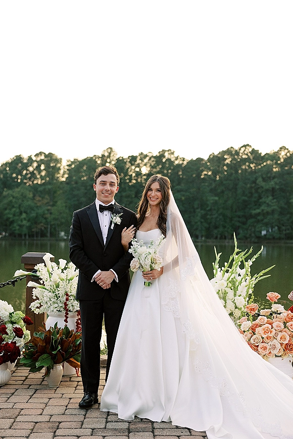 Couple portrait of bride holding a white and green bouquet beside groom in black tuxedo on a stone patio overlooking a lake