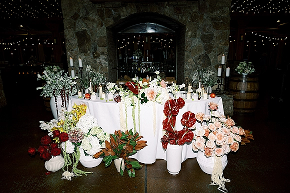 Wedding sweetheart table with sweetheart table flowers, candles, and fruit in ceramic vases, set before a stone fireplace with string lights