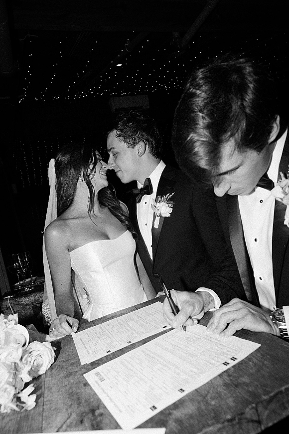 Wedding signing as bride and groom sign marriage license at a table with bouquet and wine glasses under string lights in a dark reception room