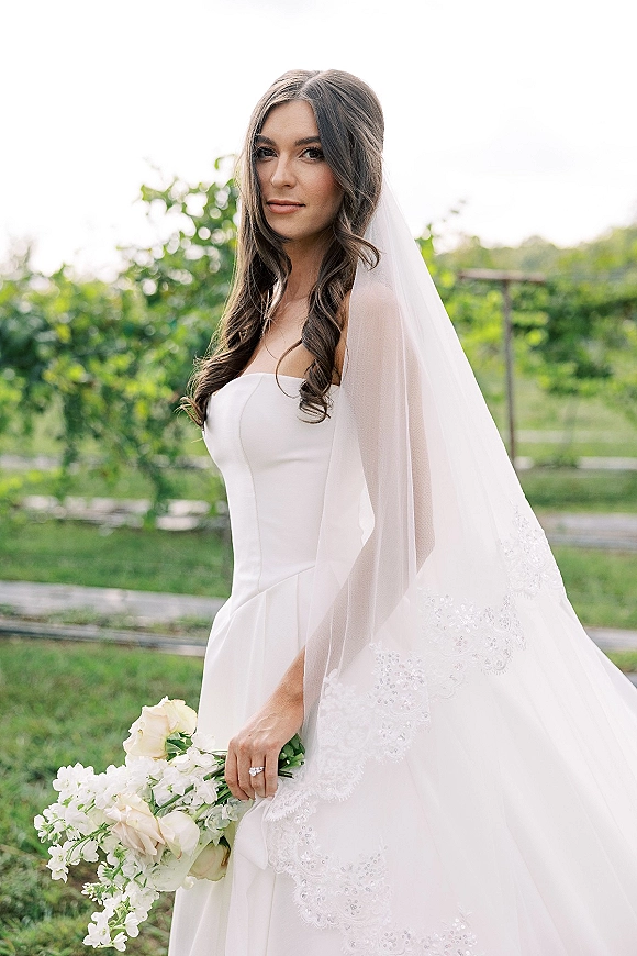 Bridal portrait of a bride holding bouquet in a strapless wedding dress, looking over her shoulder with a long veil in a garden pathway setting