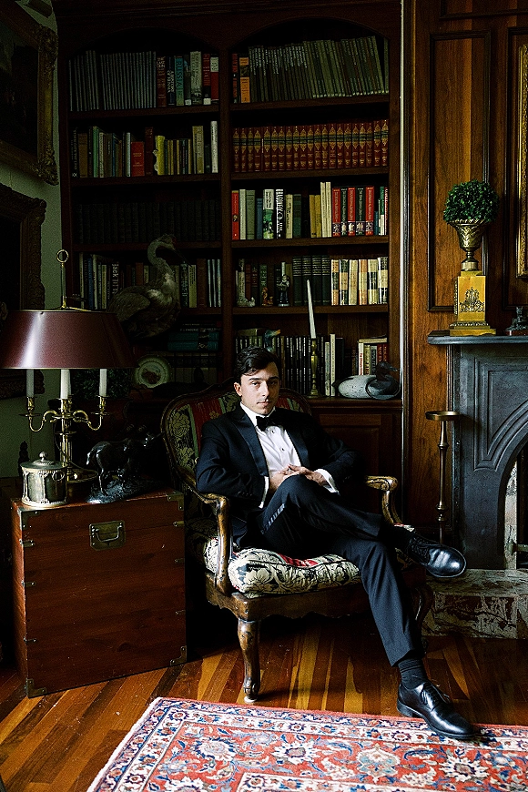 Groom portrait in black tuxedo with bow tie, seated in an upholstered armchair before library bookshelves and a fireplace mantel