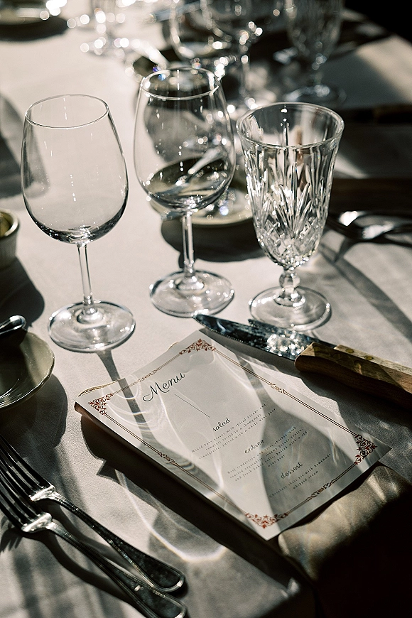 Wedding place setting with a wedding menu card, crystal goblet and wine glasses, and silver flatware on a sunlit reception table