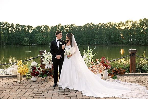 Couple portrait of bride and groom holding a bouquet, her cathedral veil flowing as they stand by a lakeside patio at sunset