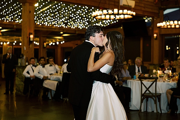 Wedding first dance kiss as bride in a strapless satin gown and groom in a tuxedo embrace under string lights in a wooden hall