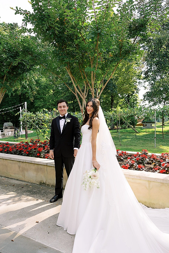 Couple portrait of bride and groom holding hands, her cathedral veil and white bouquet flowing in a string-lit garden walkway.