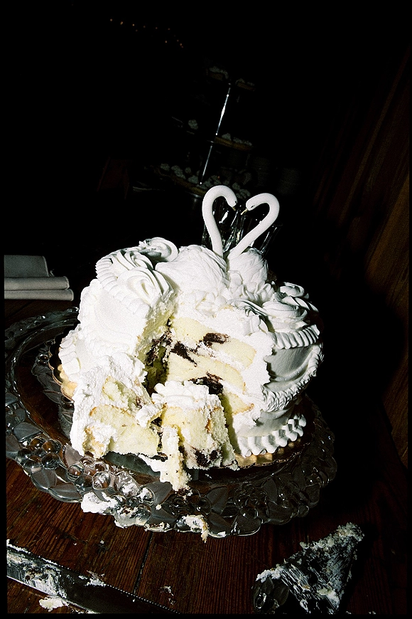 Wedding cake with swan cake topper, white frosting on a glass stand, partially cut with crumbs scattered on a wooden table in a dark room