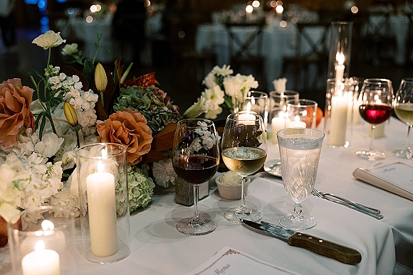 Reception tablescape with a candlelit wedding table, roses and hydrangea centerpiece, wine glasses, and tall cylinder candles under bistro lights