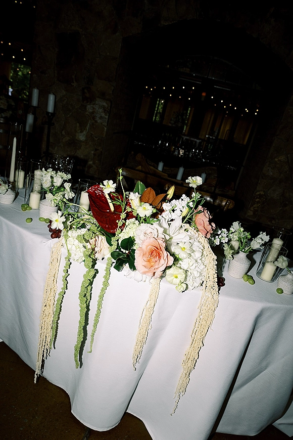 Reception tablescape with wedding table centerpiece of hanging amaranthus, candles in glass hurricanes and grapes on white linen by a stone wall