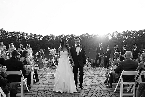 Wedding recessional as bride and groom walk up the aisle, guests clapping on a brick patio by a lake in warm sunset light