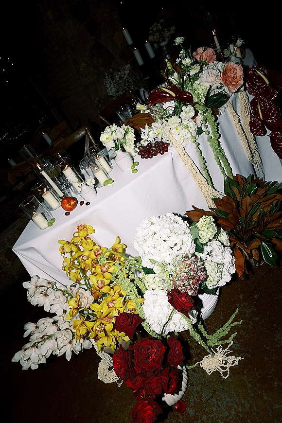 Reception tablescape with a floral table centerpiece of roses, orchids, and taper candles on white linen in a dim room with string lights