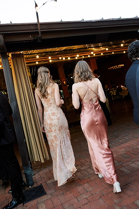 Wedding guest dresses in satin slip and floral lace as women walk a brick patio under string lights toward a draped pavilion reception