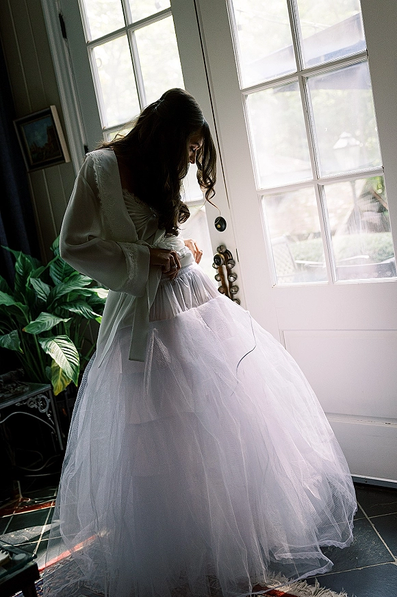 Bride getting ready in a bridal robe, adjusting a tulle wedding dress by French doors with soft window light and houseplants nearby