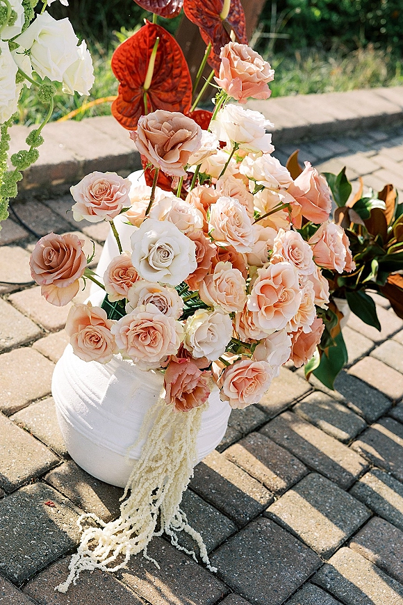 Wedding floral arrangement with peach rose centerpiece in a white ceramic urn, featuring anthurium and hanging amaranthus on a sunny patio