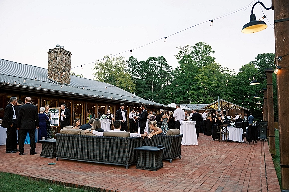 Wedding cocktail hour setup with high top tables under string lights on a brick patio courtyard by a rustic lodge and trees