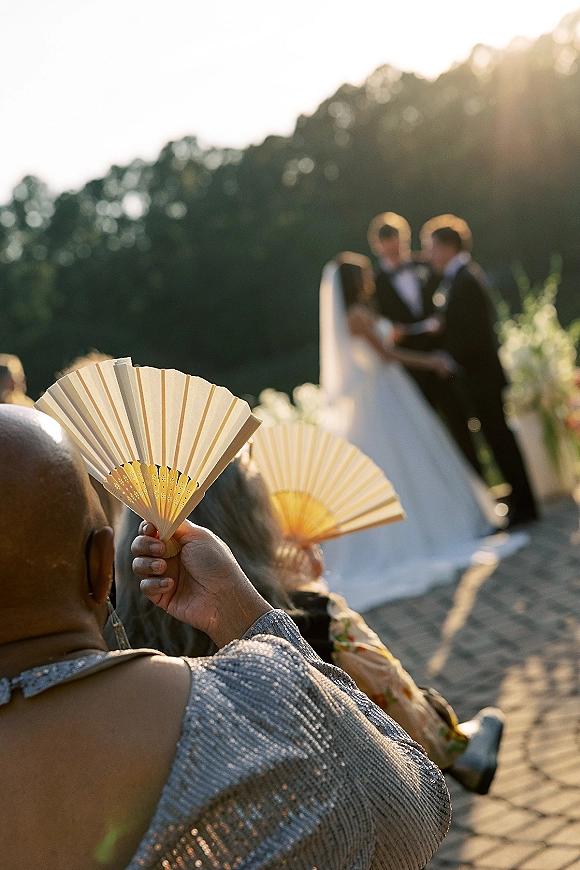 Wedding ceremony with guests holding hand fans as the veiled bride and tuxedoed groom stand on a sunlit brick walkway under trees