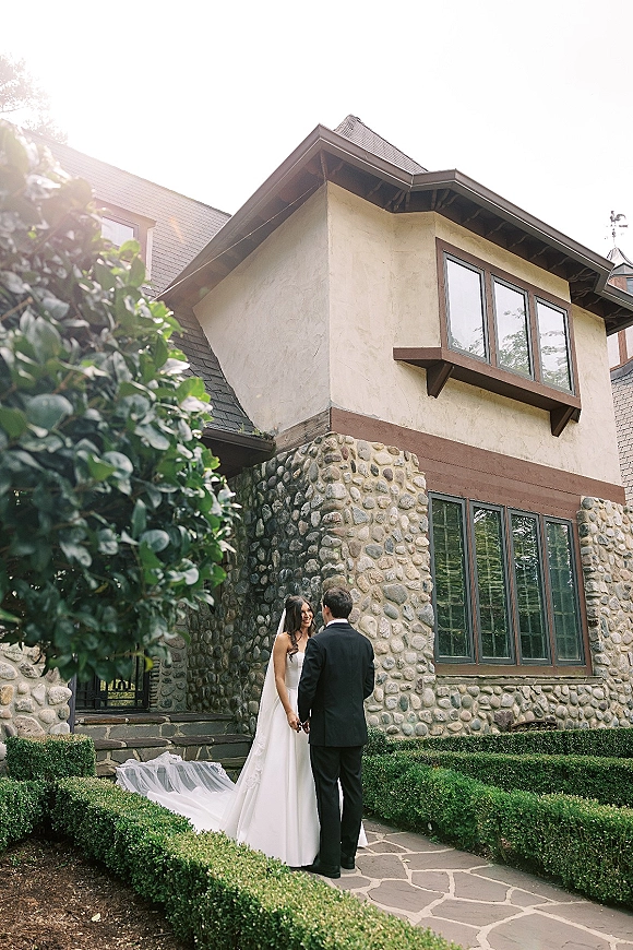 Couple portrait of bride and groom holding hands, her long veil and satin dress beside his tuxedo on a stone walkway by a windowed house