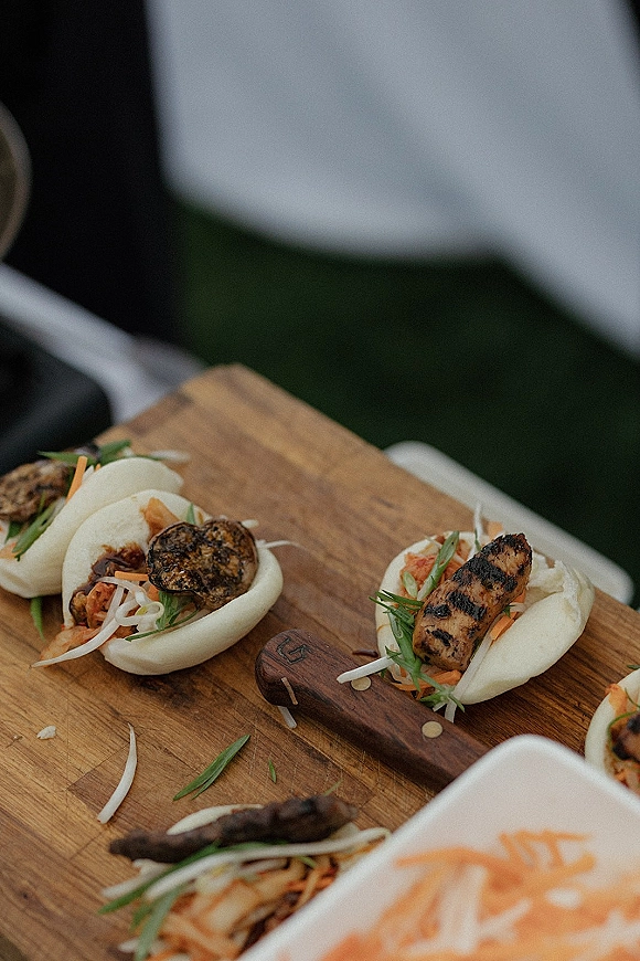 Wedding cocktail appetizers of steamed bao buns with grilled meat, shredded veggies and herbs arranged on a wood serving board outdoors