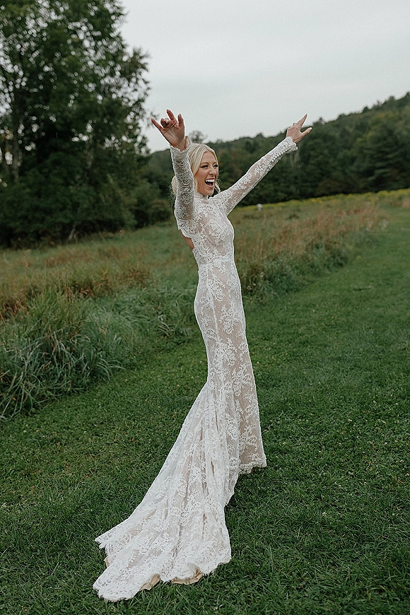 Bridal portrait of a laughing bride with arms raised in a long-sleeve lace high-neck gown, standing in a wildflower field under overcast sky