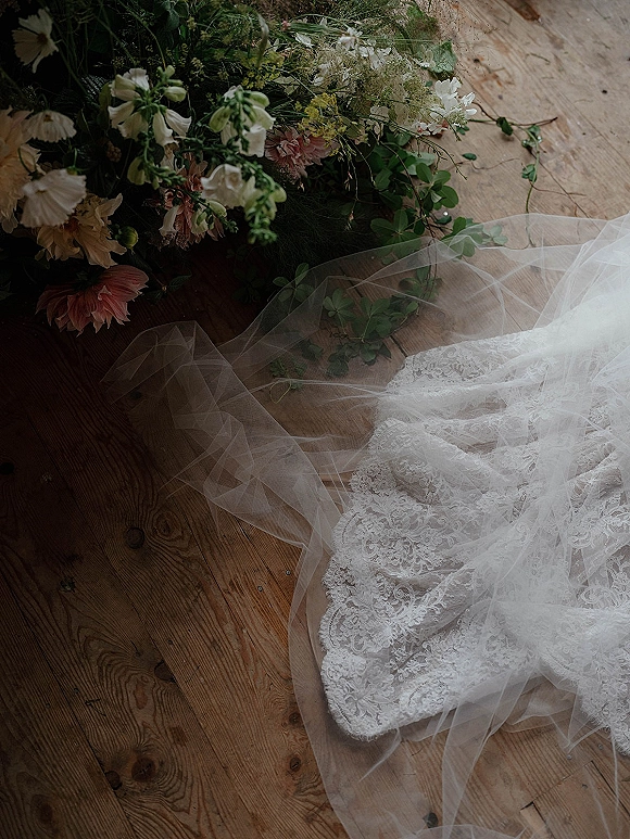 Wedding dress detail with lace wedding dress and tulle skirt laid on wood floorboards beside a wildflower bouquet and veil