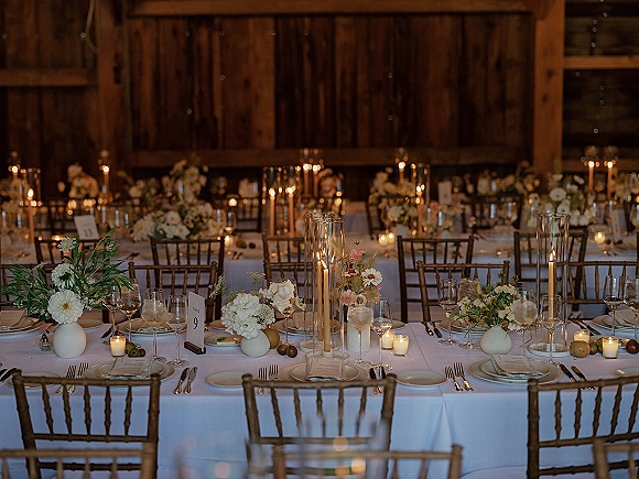 Reception tablescape with wedding table decor featuring tall hurricane candles, bud vase florals, and gold chiavari chairs in a wood barn interior