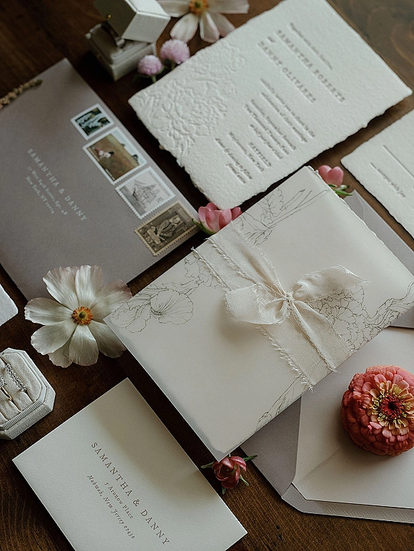 Invitation suite wedding invitation flat lay with deckle edge paper, silk ribbon bow, wax seal, rings, and flowers on a wood table