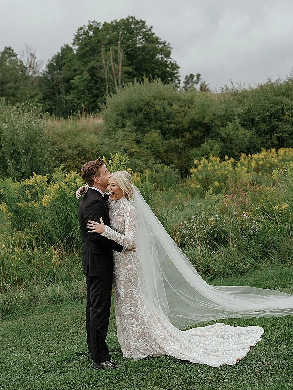 Couple portrait of bride and groom embrace, groom kissing her forehead as long veil trails in a wildflower meadow under cloudy sky