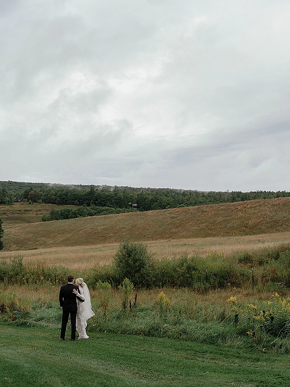 Couple portrait of bride and groom from behind embracing in a green meadow, her veil trailing under a cloudy sky and rolling hills