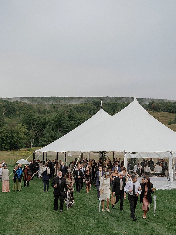 Wedding reception tent with sailcloth wedding tent and clear sidewalls as guests in formalwear hold umbrellas on a green lawn by rolling hills