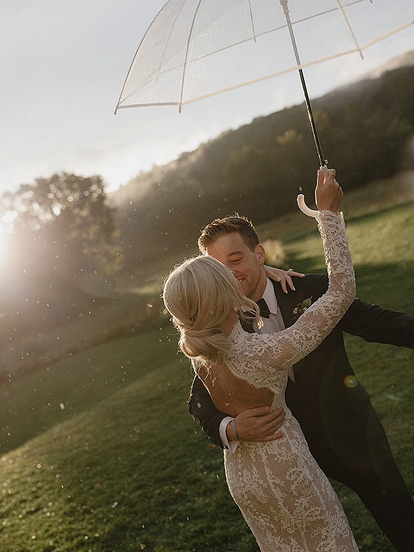 Wedding couple portrait under a clear umbrella, bride in lace long sleeves and groom in tuxedo kissing in a rainy sunset field