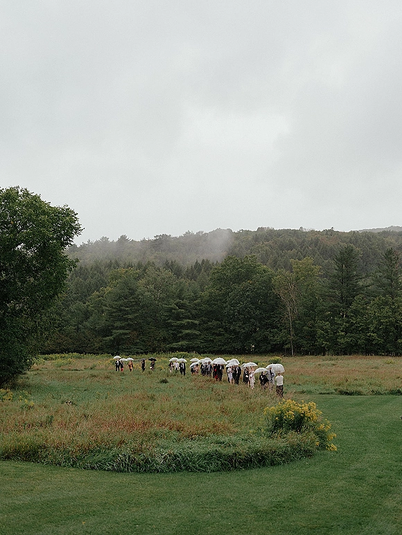Wedding processional with wedding party walking across a foggy meadow, bridesmaids carrying clear umbrellas in suits and dresses under overcast sky