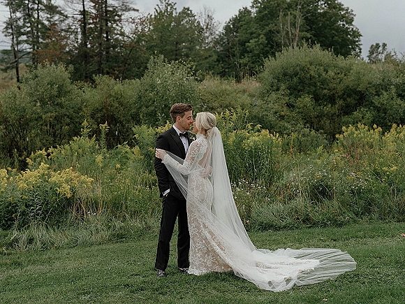 Couple portrait of bride and groom embrace, her long sleeve lace dress and cathedral veil flowing in a wildflower meadow under overcast sky